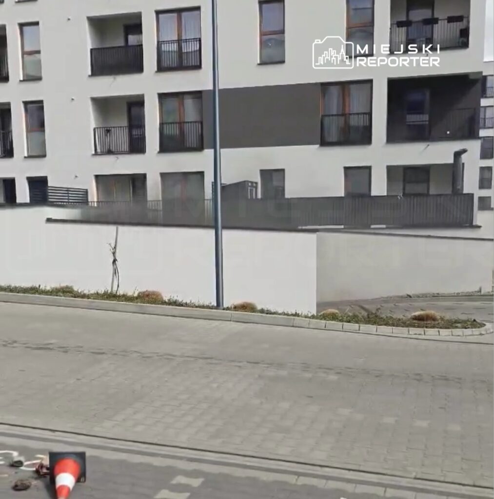 Nowoczesny budynek mieszkalny z balkonami, widok na ulicę z leżącym pachołkiem drogowym.