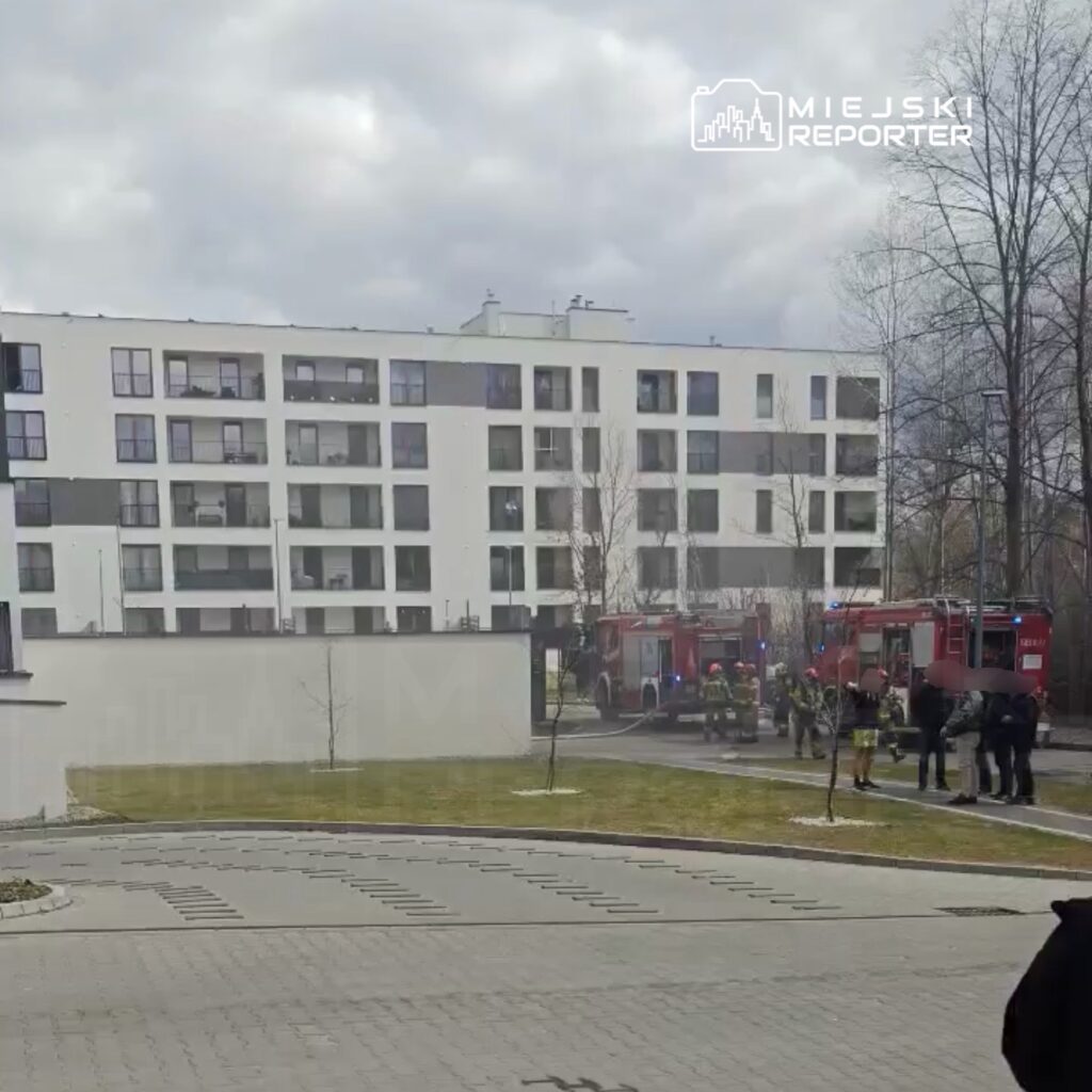 Grupa strażaków i ratowników przy wozach strażackich przed nowoczesnym budynkiem mieszkalnym w pochmurny dzień.