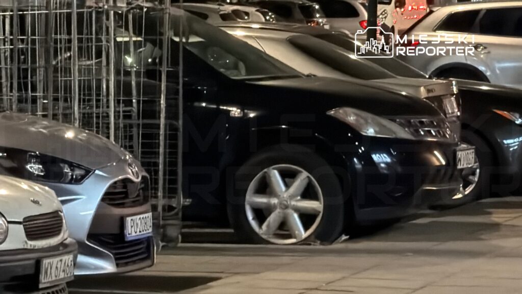 Czarny samochód zaparkowany na chodniku z jednym płaskim kołem, obok innych pojazdów i metalowych ogrodzeń.