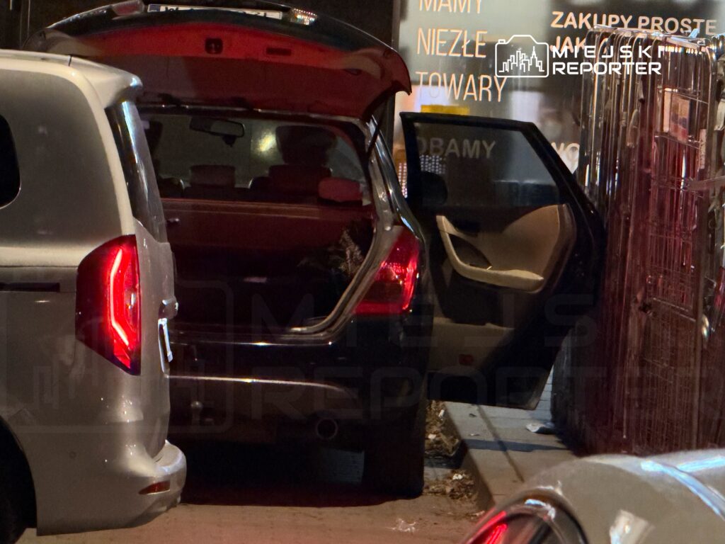 Czarny samochód z otwartą klapą bagażnika stoi obok białego vana na ulicy, w tle widoczny napis reklamowy.