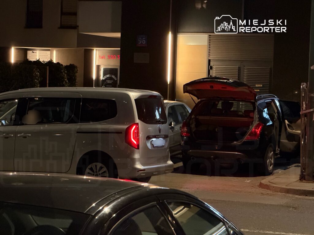 Dwa samochody zaparkowane na ulicy, jeden z otwartą klapą bagażnika, w tle oświetlony budynek z reklamą.