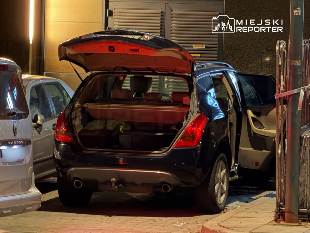 Czarny samochód z otwartym bagażnikiem stoi na parkingu, obok zaparkowany jest biały pojazd.