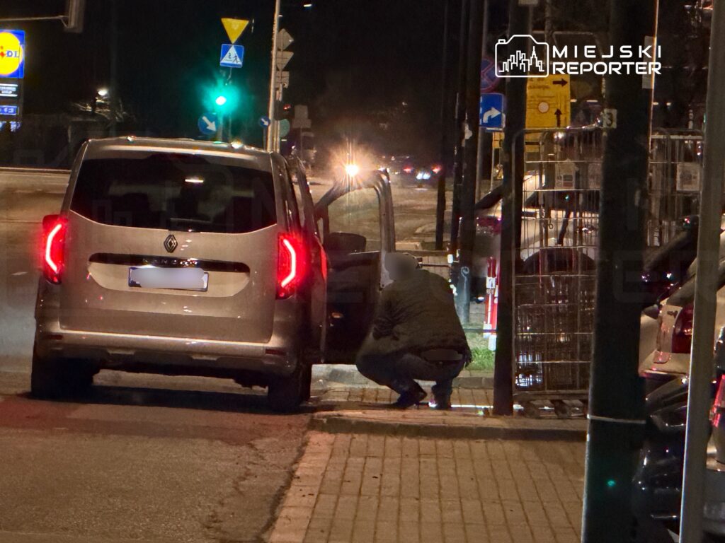 Mężczyzna w ciemnej kurtce schyla się przy otwartych drzwiach samochodu na nocnej ulicy.