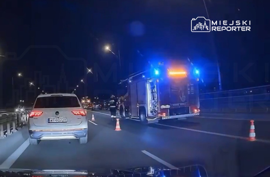 Na drodze nocą stoi wóz strażacki z włączonymi sygnałami świetlnymi, obok ustawione są pachołki.