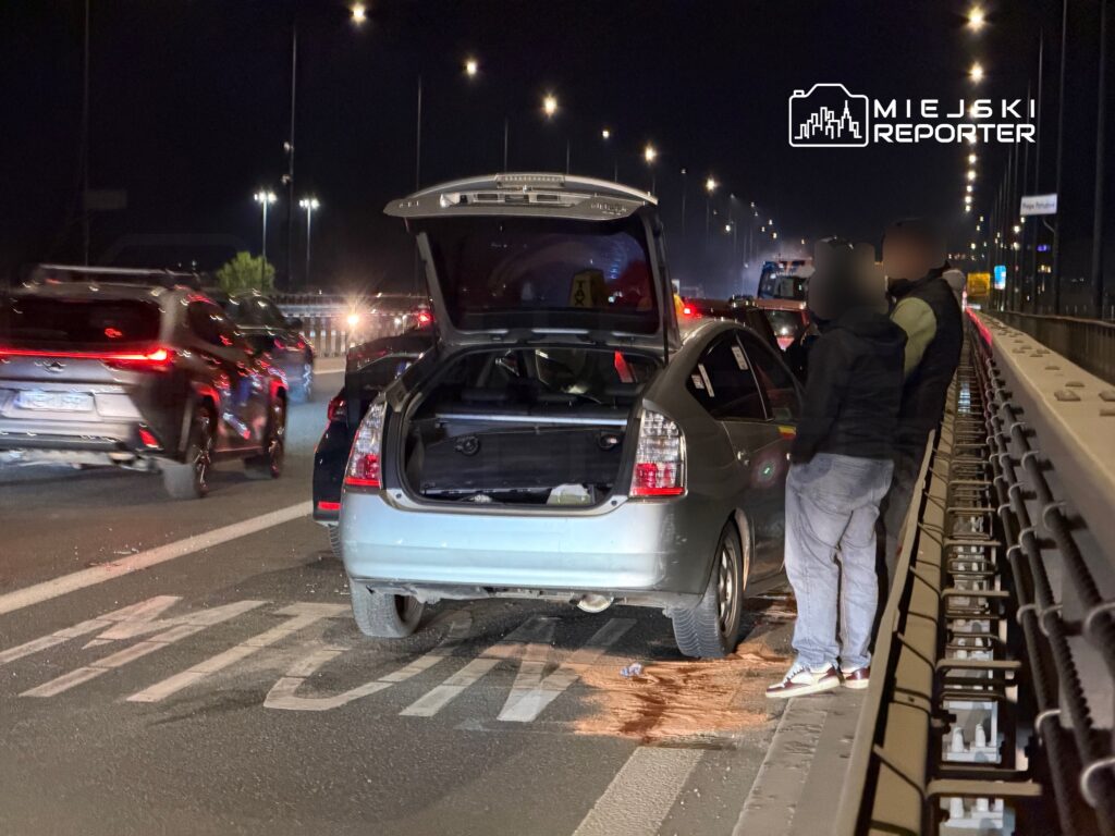 Na drodze nocą, mężczyzna przy otwartym bagażniku srebrnego samochodu, obok leżące plamy na asfalcie.