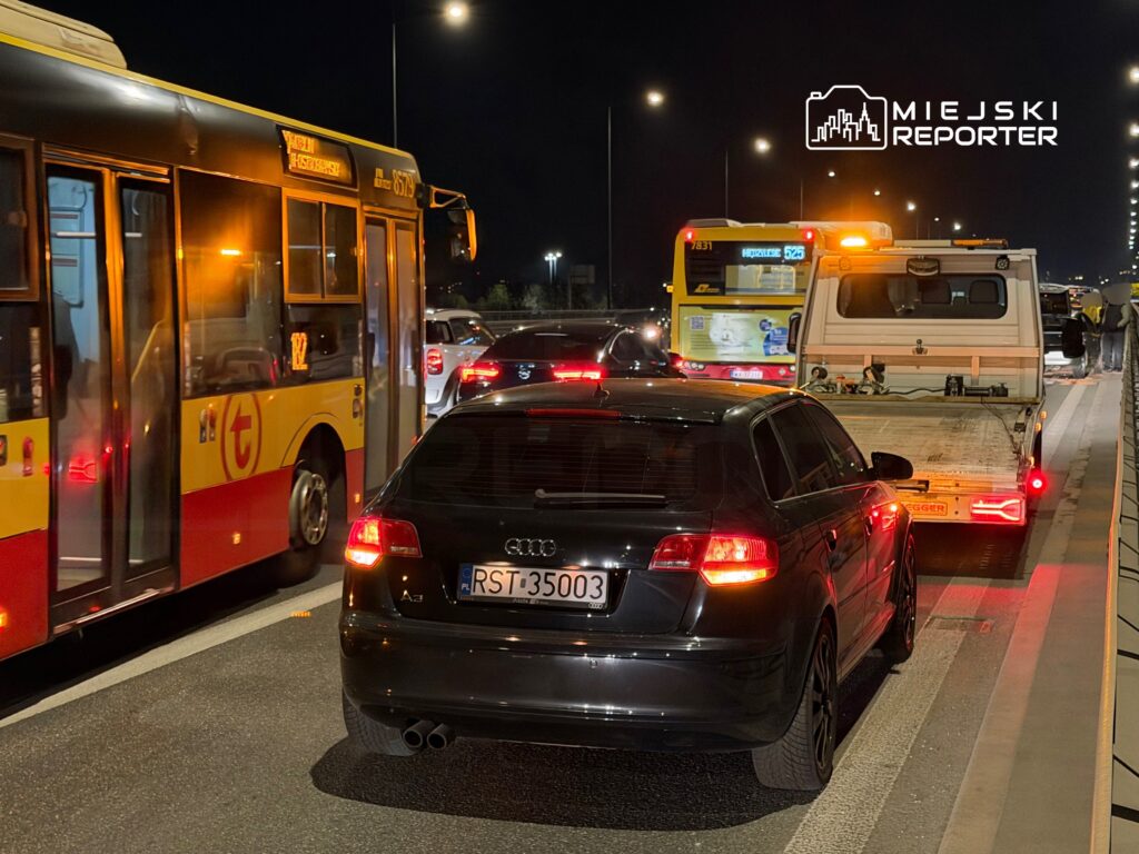 Czarny samochód osobowy zatrzymany w korku obok czerwono-żółtego autobusu nocą na miejskiej drodze.