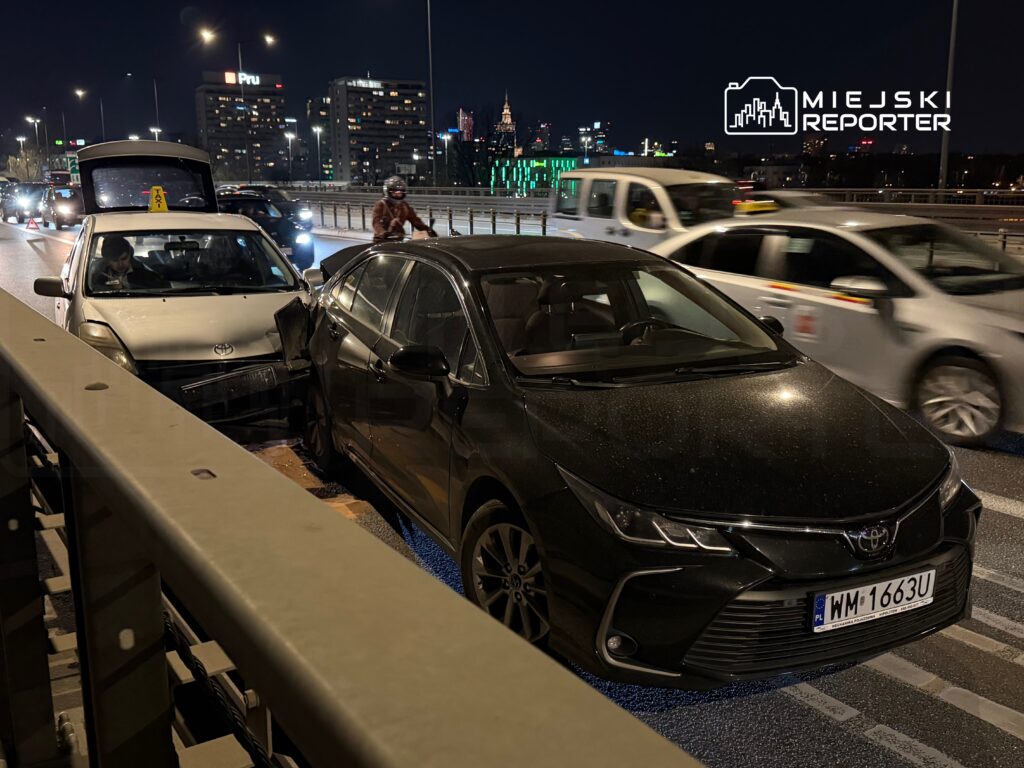 Dwa samochody osobowe zderzone na ruchliwej ulicy nocą, w tle widać miejskie budynki i światła.