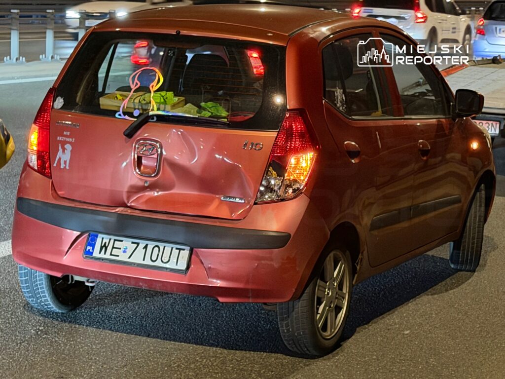 Czerwony samochód osobowy z uszkodzoną tylną częścią stoi na drodze, obok widać inne pojazdy.