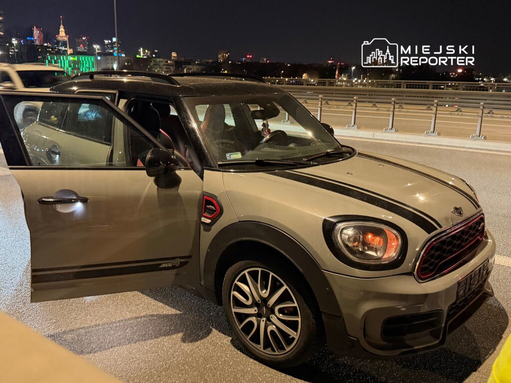 Srebrny samochód osobowy z otwartymi drzwiami stoi na nocnej drodze, w tle widoczne oświetlone wieżowce miasta.