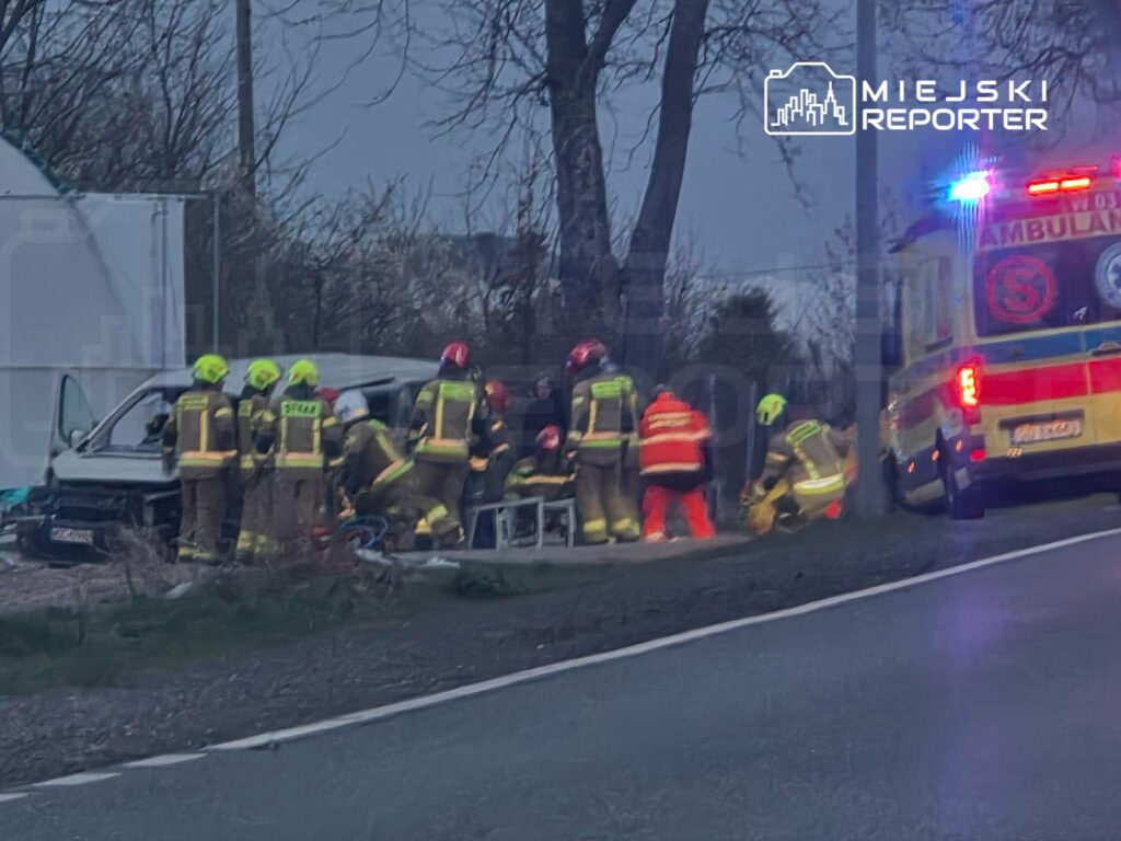 Grupa strażaków w kaskach pomaga osobom poszkodowanym w wypadku drogowym obok uszkodzonego pojazdu.