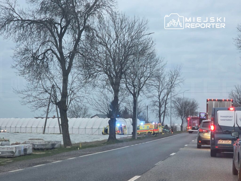 Na drodze stoi zespół ratunkowy oraz straż pożarna, obok białe tunele foliowe, a w tle widać zator drogowy.