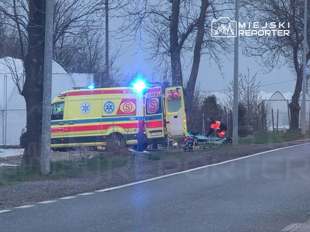 Karetka pogotowia z włączonymi sygnałami świetlnymi zatrzymana przy drodze, a ratownik przygotowuje nosze.