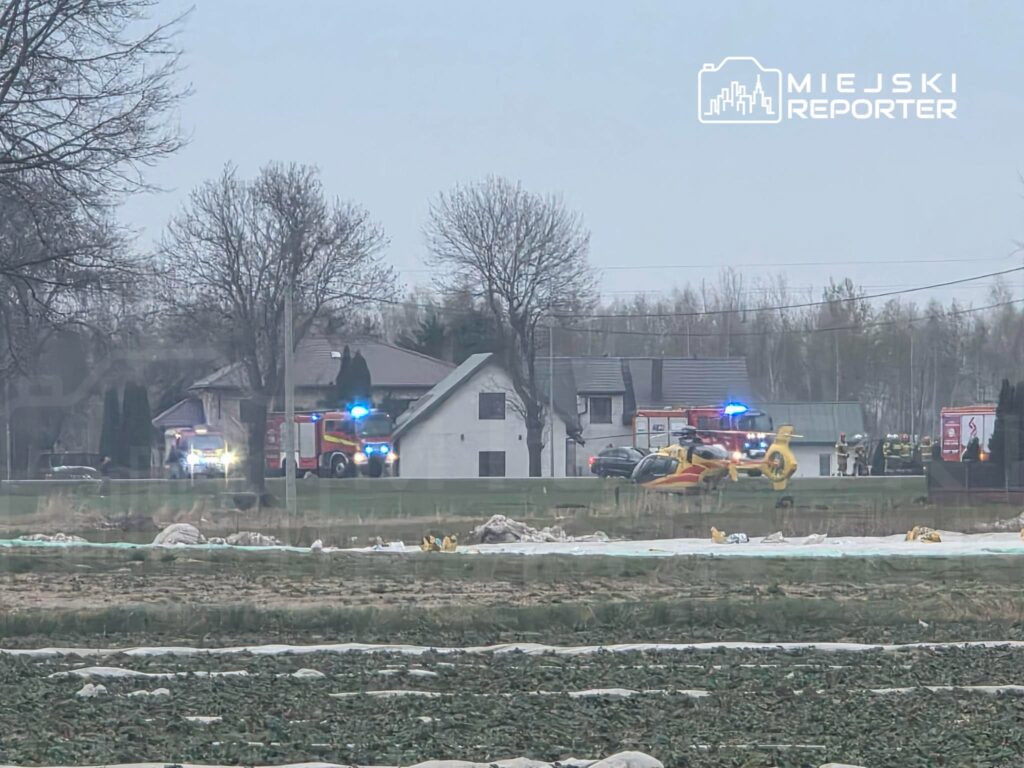 Na polu znajdują się pojazdy ratunkowe oraz helikopter, w otoczeniu domów i drzew. Akcja ratunkowa w toku.