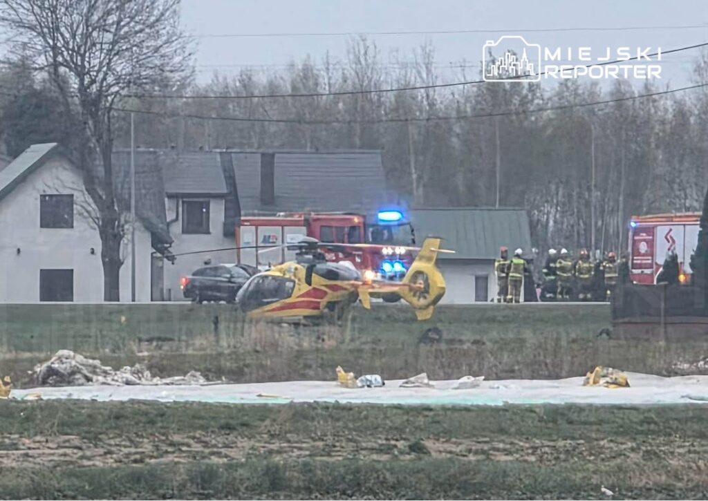 Helikopter ratunkowy lądował na polu, obok grupy strażaków i pojazdów ratunkowych, w otoczeniu domów.