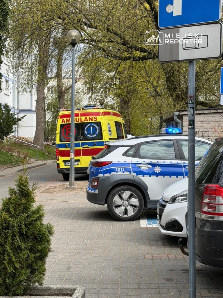 Karetka pogotowia oraz radiowóz policyjny zaparkowane na ulicy w otoczeniu drzew i budynków mieszkalnych.