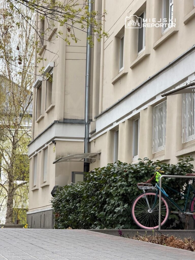 Różnokolorowy rower z różowymi kołami oparty o metalowy płot, w tle widoczna elewacja budynku mieszkalnego.