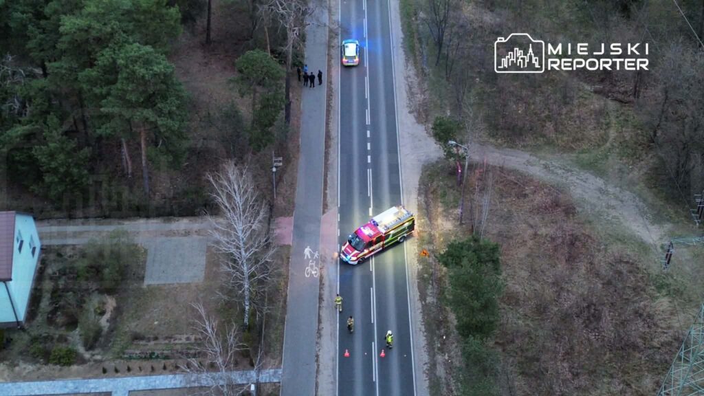 Na drodze w lesie stoi wóz strażacki z włączonymi sygnałami świetlnymi, obok kilku ratowników oraz radiowóz policyjny.