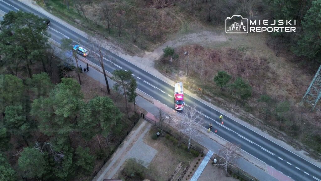 Straż pożarna z włączonymi sygnałami świetlnymi stoi na poboczu drogi, obok grupy osób w kamizelkach odblaskowych.