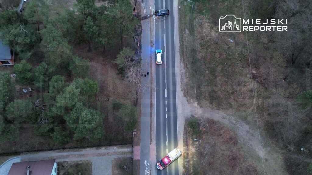 Na drodze znajduje się nieoznakowany radiowóz oraz karetka, obok leży uszkodzony samochód. W tle drzewa i pusta przestrzeń.