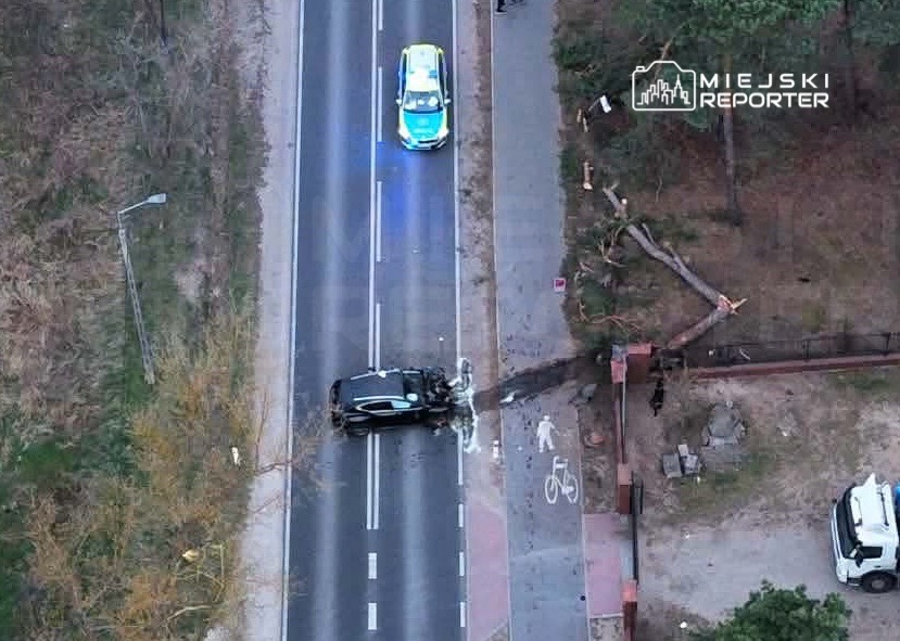Na drodze leży uszkodzony samochód, obok stoi radiowóz policyjny z włączonymi sygnałami świetlnymi.