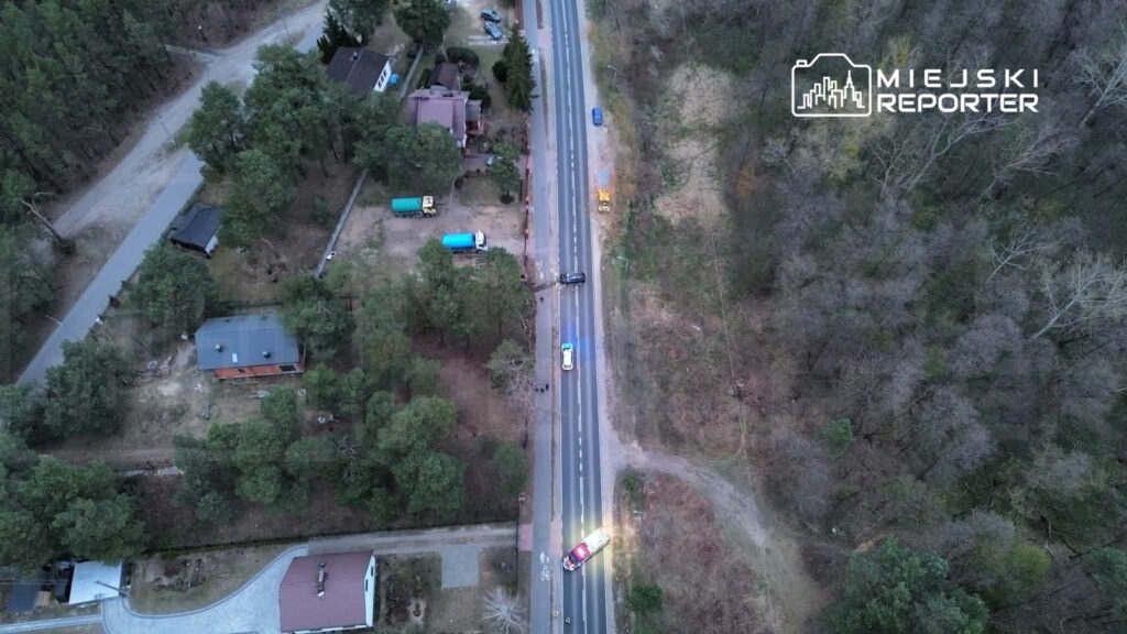 Pojazdy służb ratunkowych zatrzymane przy drodze, otoczone przez las i domy jednorodzinne w okolicy.