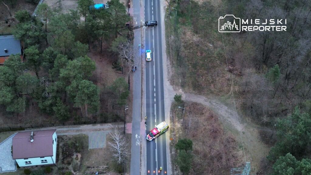 Na drodze w lesie stoi ambulans, a obok niego zatrzymane są dwa pojazdy. W tle widoczni są przechodnie.