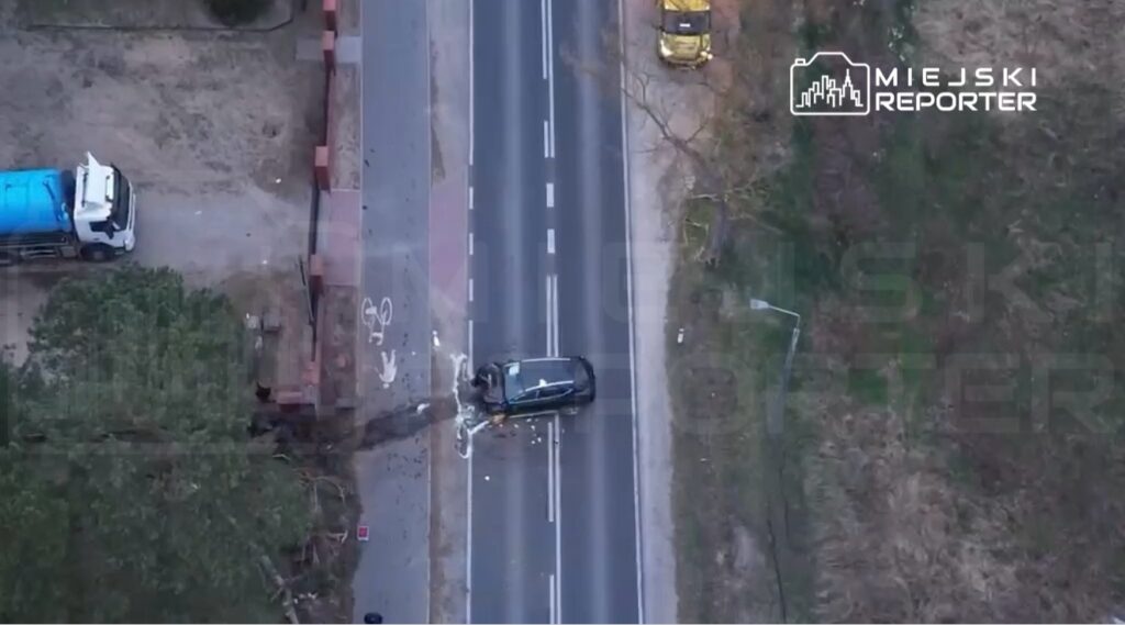 Czarny samochód osobowy leży na asfalcie drogi, obok niego widać uszkodzenia i pozostałości po wypadku.