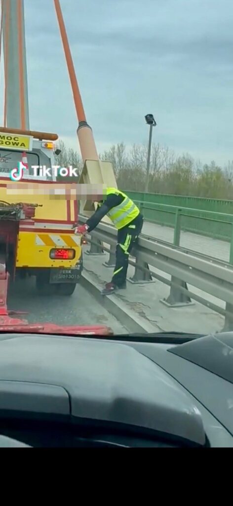 Mężczyzna w odblaskowej kamizelce naprawia tył lawety na moście, obok barierki ochronnej.