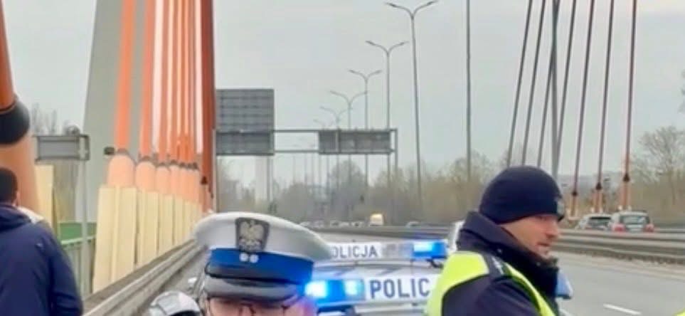 Policjanci w mundurach kontrolują ruch na moście, obok radiowóz z włączonymi sygnałami świetlnymi.