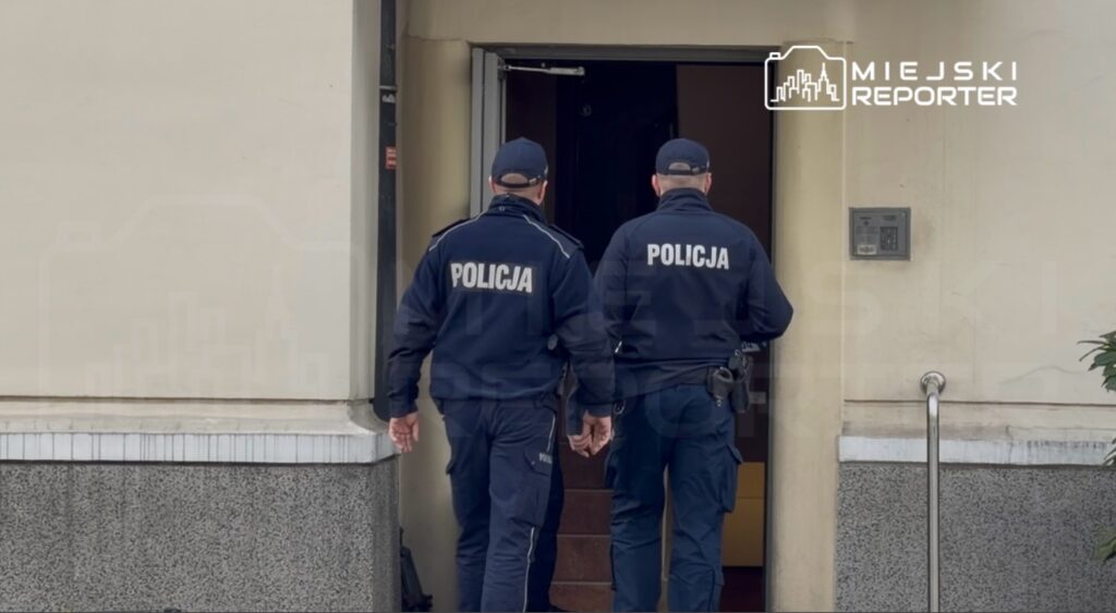 Dwóch policjantów w mundurach wchodzi do budynku przez otwarte drzwi.