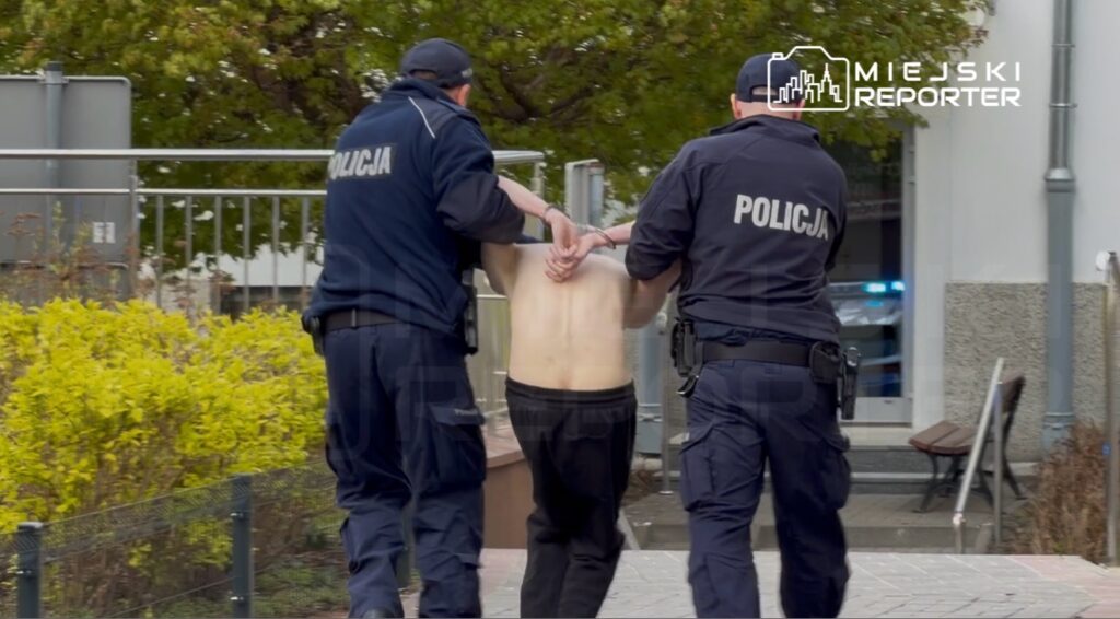 Dwaj policjanci prowadzą mężczyznę bez koszuli, trzymając go za ręce, na tle miejskiego otoczenia.