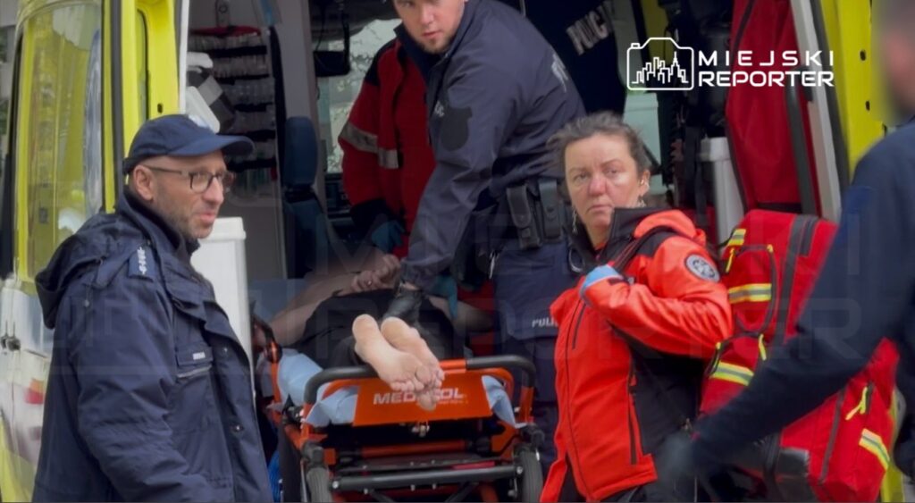 Ratownicy medyczni transportują pacjenta na noszach do karetki, a obok nich stoi policjant i kobieta w czerwonym uniformie.