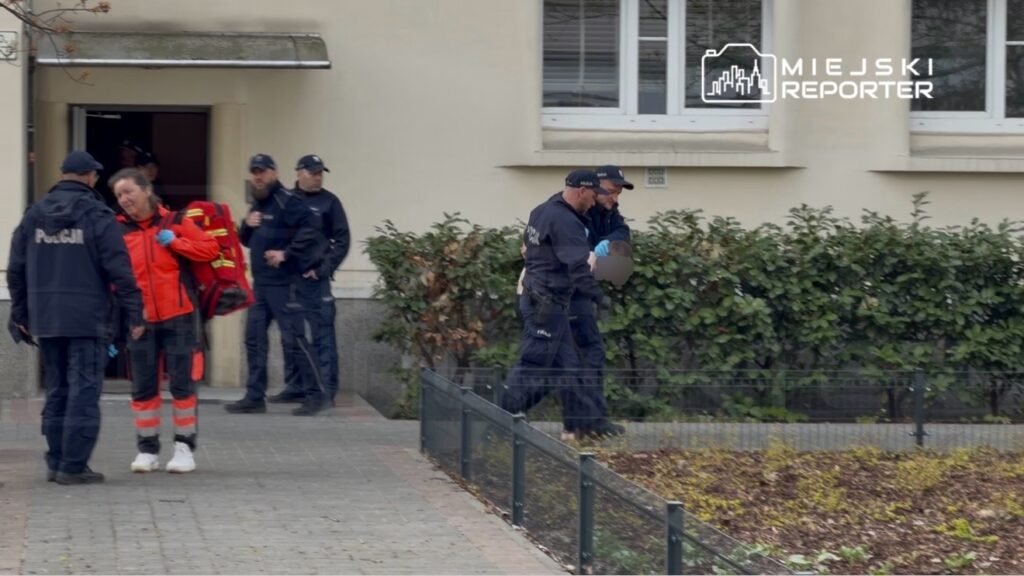 Kobieta w pomarańczowym stroju ratownika medycznego rozmawia z policjantem, podczas gdy inni funkcjonariusze obserwują sytuację.