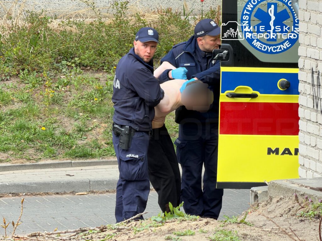 Dwóch policjantów transportuje osobę bez koszuli w kierunku karetki pogotowia na ulicy.