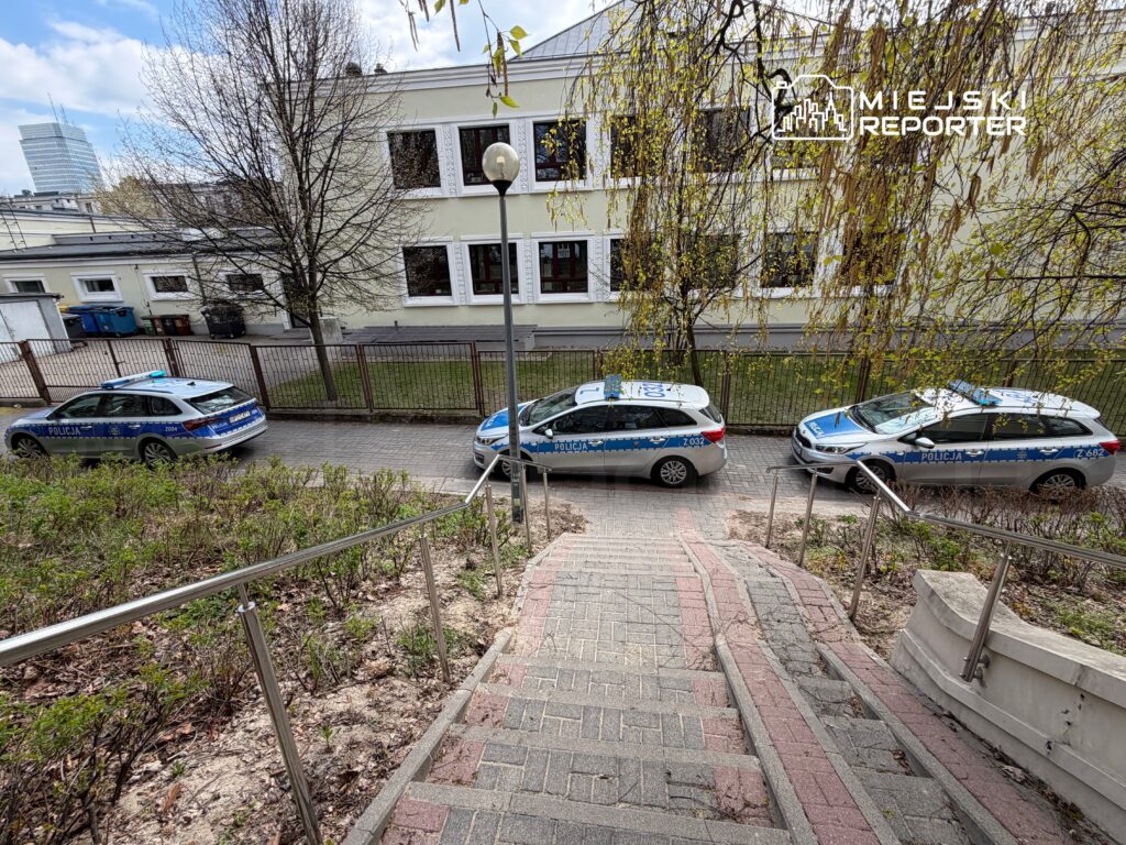 Trzy radiowozy policyjne zaparkowane wzdłuż chodnika obok budynku, otoczone zielenią i drzewami.