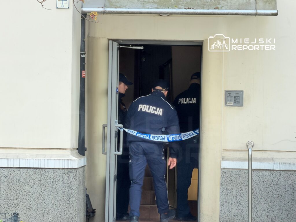 Trzech policjantów w mundurach wchodzi do budynku, przechodząc przez otwarte drzwi z taśmą policyjną.