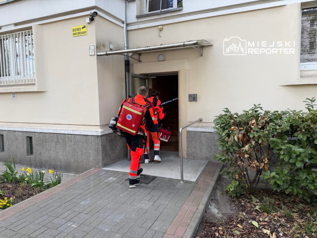 Dwóch ratowników medycznych w pomarańczowych uniformach wchodzi do budynku z monitoringiem.