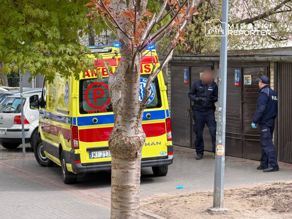 Zespół ratunkowy w karetce interweniuje w pobliżu zamkniętego budynku, a policjanci prowadzą rozmowę.
