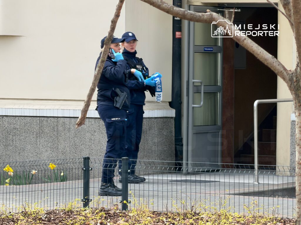Dwie policjantki rozmawiają na zewnątrz budynku, jedna trzyma telefon, obie w mundurach i rękawiczkach.