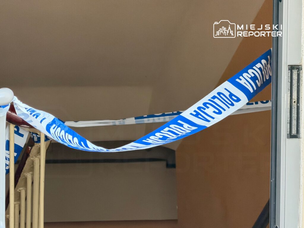 Nieprzechodnia taśma policyjna zamknięta w wejściu do budynku, z widoczną klatką schodową w tle.