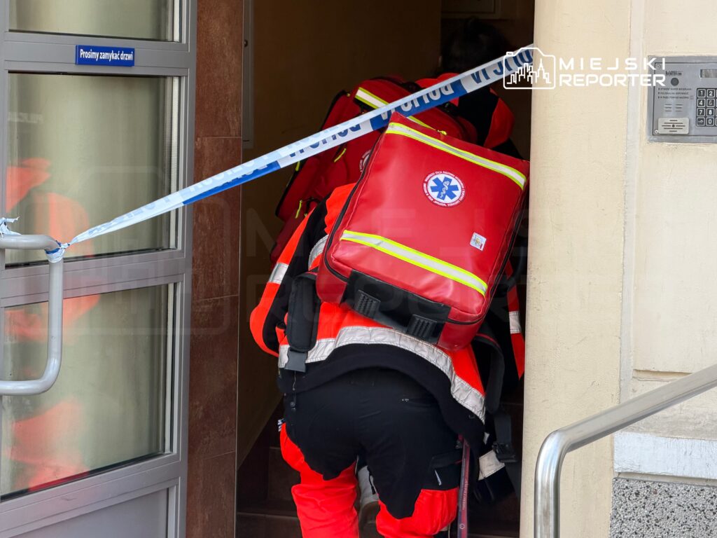 Ratownicy medyczni w czerwonych uniformach wchodzą do budynku, przechodząc przez zaklejoną taśmą drzwi.