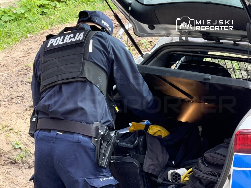 Funkcjonariusz policji przeszukuje bagażnik radiowozu w otoczeniu zieleni.