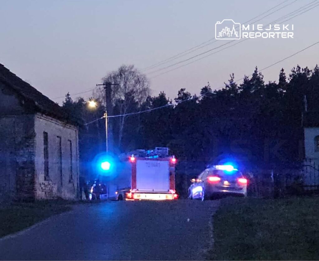 Strażacki wóz z niebieskimi światłami oraz radiowóz policji na drodze w pobliżu opuszczonego budynku.