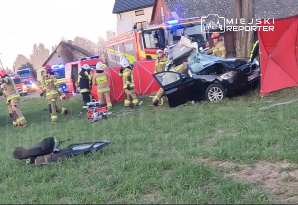 Grupa strażaków pracuje przy wypadku samochodowym, obok uszkodzonego czarnego auta, w otoczeniu sprzętu ratunkowego.