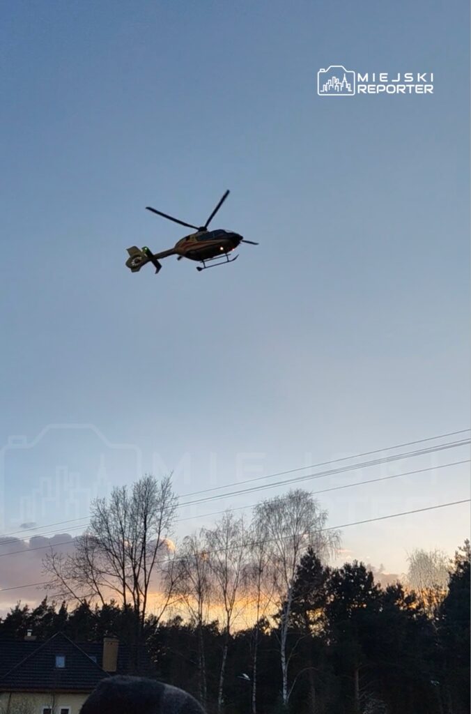 Śmigłowiec ratunkowy przelatuje nad osiedlem, w tle widoczne drzewa i zachodzące słońce.