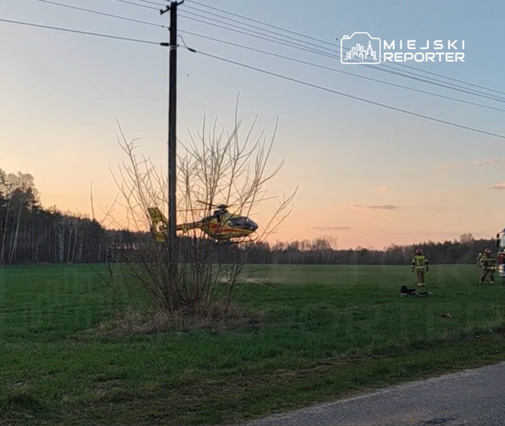 Śmigłowiec ratunkowy ląduje na zielonej łące, obok grupy strażaków w uniformach.