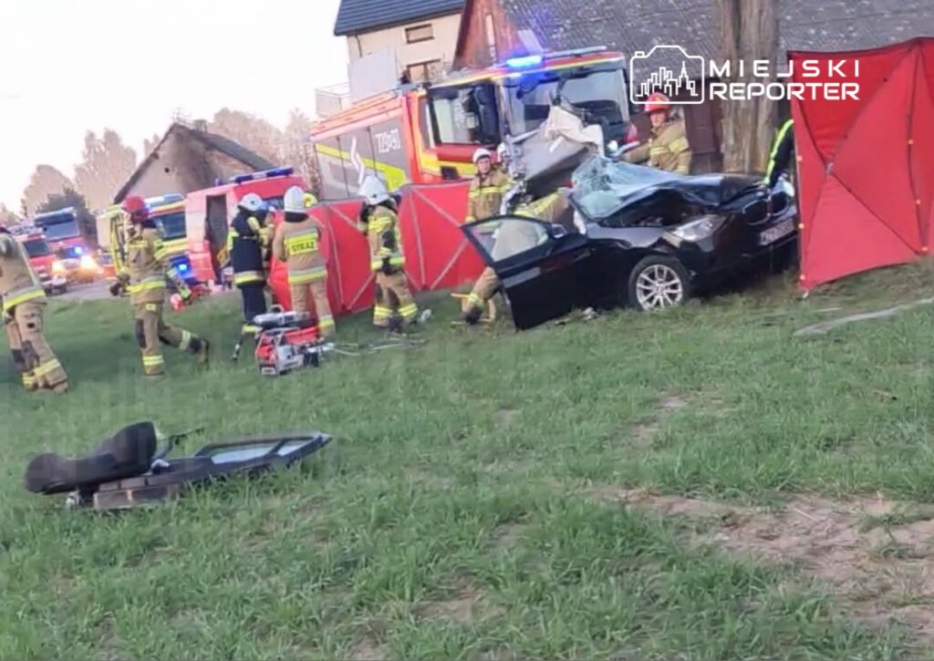 Grupa strażaków w mundurach pracuje przy rozbitym czarnym samochodzie, otoczona przez pojazdy ratunkowe.