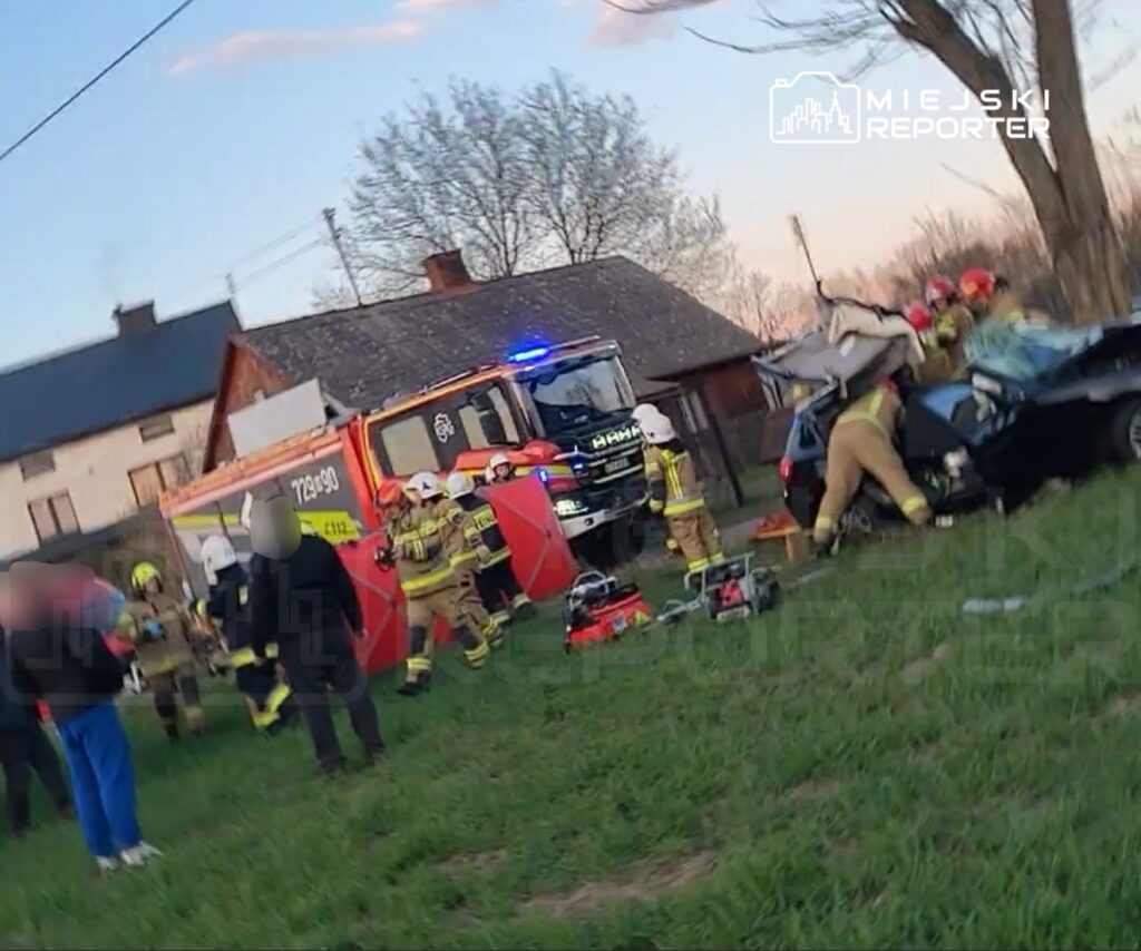 Strażacy pracują nad rozbitym samochodem osobowym w terenie, obok widoczny wóz strażacki z włączonymi sygnałami.