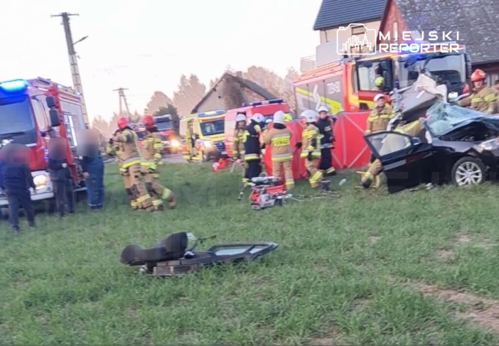 Grupa strażaków w mundurach ratunkowych pracuje przy zniszczonym samochodzie na łące, obok pojazdów ratunkowych.