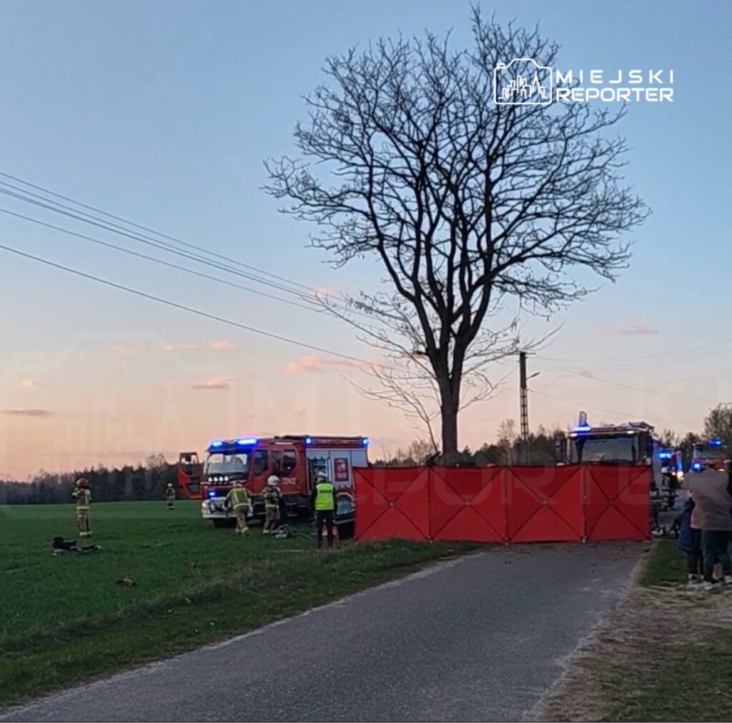 Strażacy pracują przy wozach strażackich na polu, otoczeni przez czerwoną barierkę.